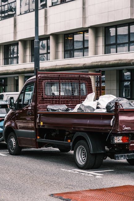 The image displays a large collection of discarded rubbish piled around two waste bins situated on a paved pavement area, with an urban commercial background that includes a parked silver car on the l